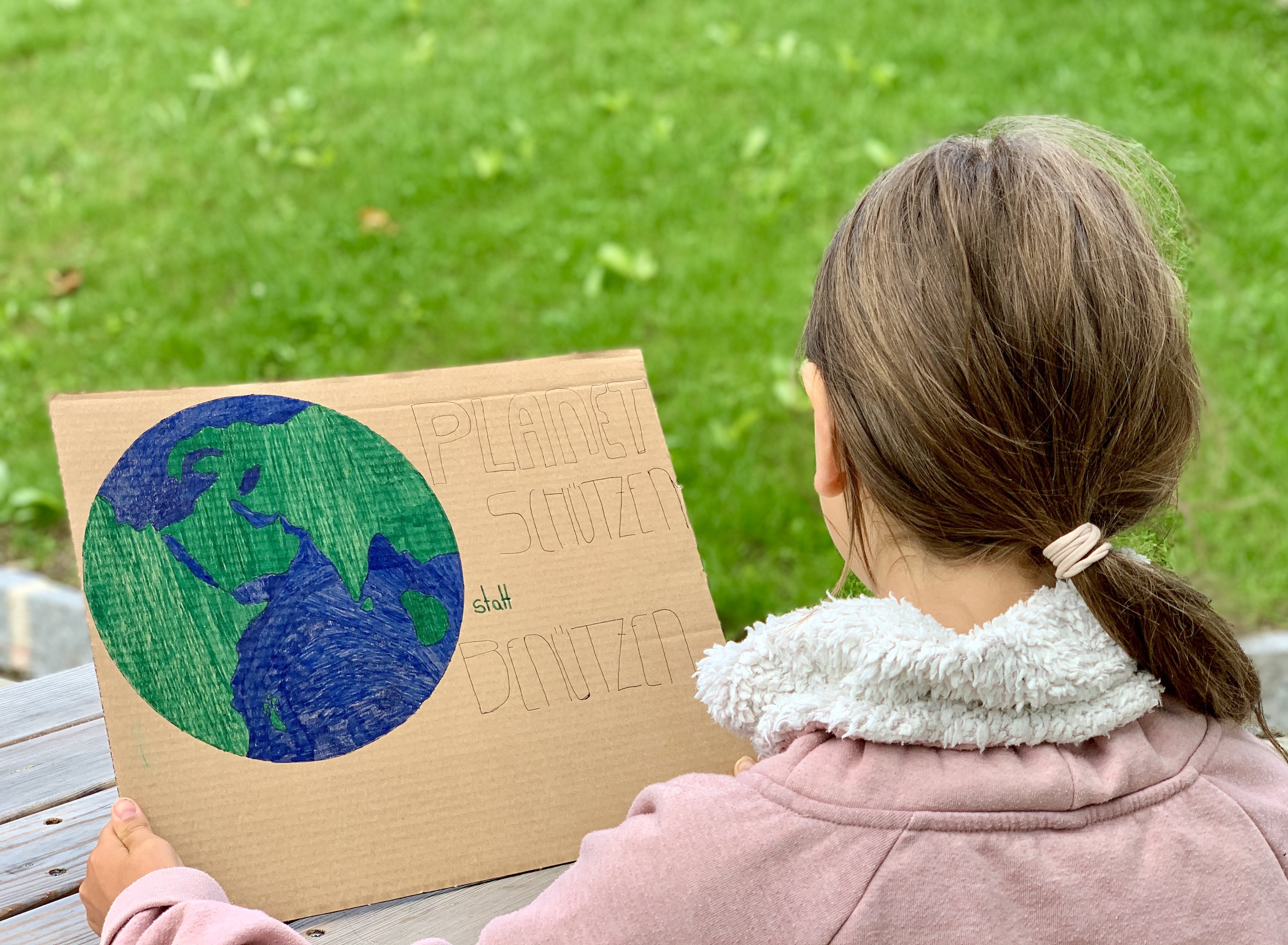 Kinderwoche 2021 – Bericht Ein Kind mit langen Haaren ist von hinten zu sehen und betrachtet ein Demo-Plakat mit einer Zeichnung der Erde und der Aufschrift: "Planeten schützen statt benützen!"